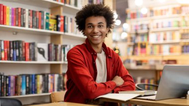 Üniversite kütüphanesinde masasında oturmuş kameraya gülümseyen Afro-Amerikan bir öğrencinin portresi.