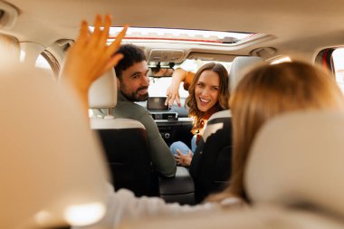 Ailenin arabada oturması, ebeveynlerin kızına dönmesi, gülümsemesi, birlikte araba yolculuğunun tadını çıkarması.