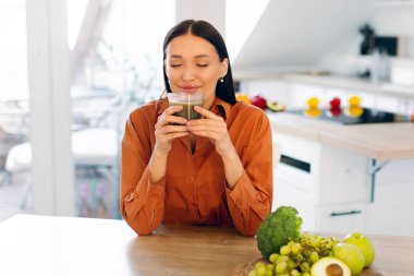 Tatmin olmuş bir kadın cam bardaktan taze sıkılmış smoothie kokan, masada yeşil meyve ve sebzelerle oturan,