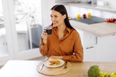 Mutlu bayanın atıştırması, kolayla bardak tutması ve fast food dağıtımından lezzetli hamburger yemesi, mutfakta oturan kadın.