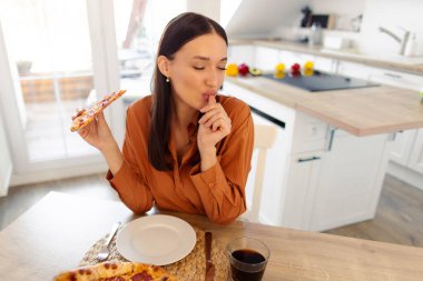 Mutlu bayan zevkle pizza yemekten ve parmaklarını kapalı gözlerle yalamaktan hoşlanıyor, mutfak masasında oturuyor, manzaranın üstünde, bedava uzay.