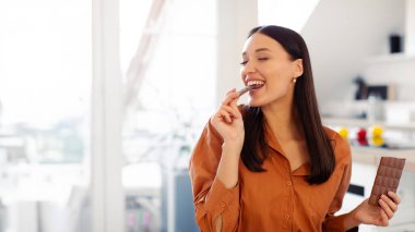 Tatmin olmuş genç bir bayan elinde fayansla siyah çikolata yiyor, tatlı tatlının tadını çıkarıyor, evde mutfakta oturuyor.