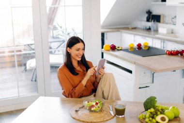 Kadın cep telefonuyla sosyal medyada gezinirken taze salatadan zevk alıyor. Parlak mutfak içlerinde masada oturuyor. Boş yer var.