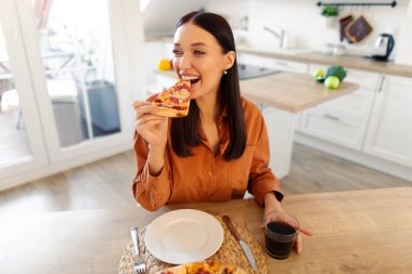 Mutlu bir kadının pizza yemesi, mutfakta masada oturması, evde aldatma yemeğinin tadını çıkarması. Abur cubur konsepti