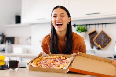 Yemek teslimatı konsepti. Çok mutlu bir kadın, elinde lezzetli pizzayla bir kutu tutuyor. Mutfağın içinde duruyor. Mükemmel bir teslimat hizmeti veriyor.