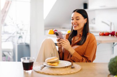 Mutfak masasında oturan rahat bir bayan, cep telefonunu kullanarak yemeği hamburgerle aldatıyor.