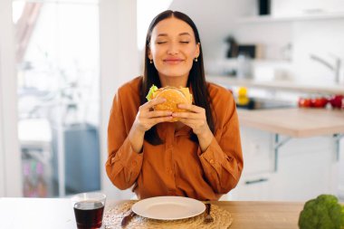 Gözleri kapalı genç bir kadın iştah açıcı lezzetli hamburgerin tadını çıkarıyor, mutfakta oturuyor ve lezzetli hamburgerler yiyor.
