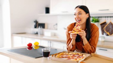 Ellerinde lezzetli bir dilim pizzayla çok heyecanlı genç bir kadın. Kokusunun ve tadının tadını çıkarıyor. Mutfakta duruyor ve boş alana bakıyor.