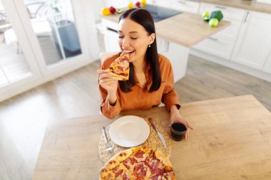 Tatmin olmuş bir bayan taze pizzayı seviyor, elinde tutuyor ve ısırıyor, mutfak masasında oturuyor, manzaranın üstünde. Abur cubur aşığı, evde aldatan yemek.