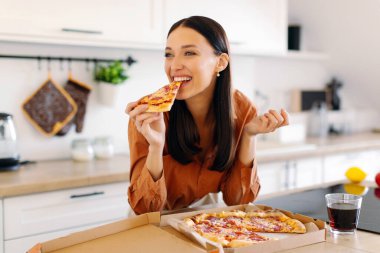 Neşeli bayan tek başına pizza yiyor, dilimliyor ve tadını çıkarıyor, yemek dağıtımından memnun, evde mutfak tezgahında duruyor.
