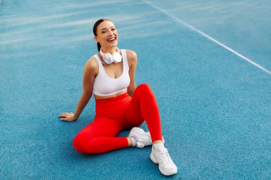 Heyecanlı Avrupalı sporcu kadın, kulaklıkları boynunda, dinleniyor, kameraya poz veriyor ve gülümsüyor, sahada oturuyor, dışarıda antremandan sonra mola veriyor.