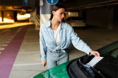 Üzgün kadın sürücü park cezasına bakıyor ya da iyi, alışveriş merkezinin yeraltı otoparkında arabasının yanında duruyor, boş yer var.