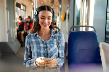 Modern tramvayda oturan mutlu bir bayan, kablosuz kulaklık takıyor ve cep telefonu kullanıyor, çevrimiçi müzik dinliyor, boş yer arıyor. Rahat şehir ulaşımı kavramı