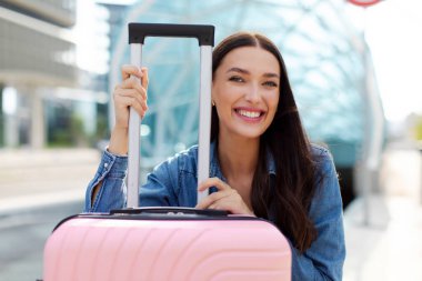 Bavulun yanında oturan mutlu bayan, yolculuk için heyecanlı, yolculuğa hazır, uçağa binmeyi bekliyor ve kameraya gülümsüyor.