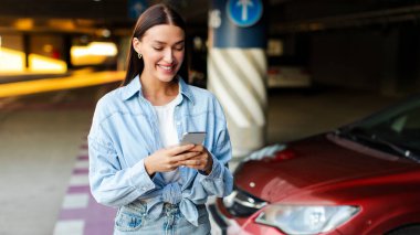 Mutlu bayan cep telefonu kullanıyor, otoparkta arabanın yanında duruyor, iletişim kuruyor ya da online uygulama kullanıyor. Araba paylaşımı, kiralama servisi veya taksi uygulaması