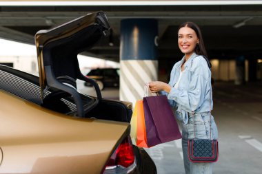 Açık araba bagajının yanında duran ve alışveriş merkezinde başarılı bir şekilde alışveriş yaptıktan sonra paketlemek için kese kağıdı tutan neşeli genç bayan.