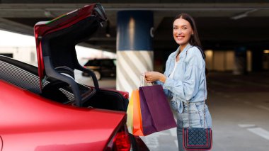 Açık araba bagajının yanında duran ve alışveriş merkezinde başarılı bir şekilde alışveriş yaptıktan sonra paketlemek için kese kağıdı tutan neşeli genç bayan.