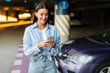 Mutlu bayan cep telefonu kullanıyor, otoparkta arabanın yanında duruyor, iletişim kuruyor ya da online uygulama kullanıyor. Araba paylaşımı, kiralama servisi veya taksi uygulaması