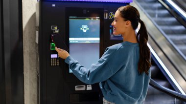 Yeraltı otoparkına para ödeyen ya da elektronik self-servis kabinini kullanarak metro ya da tren bileti alan genç bir kadın.