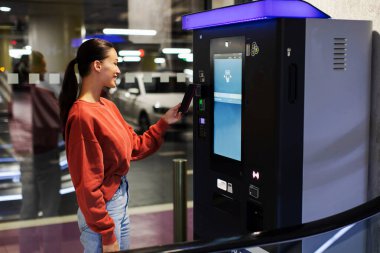 Genç bir kadın, akıllı telefonuyla park yeri ödeme makinesine ulaşamıyor. Arabasını alışveriş merkezindeki yeraltı otoparkına park etmiş.