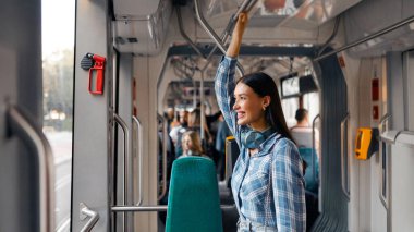 Modern tramvayda ayakta duran mutlu kadın yolcu toplu taşımada rahat bir yolculuk yapıyor, pencereye bakıyor ve gülümsüyor.