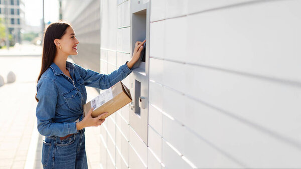 Концепция почты. Молодая женщина с коробкой на открытом автоматизированном участке машины выбирая работу на сенсорном экране, вид сбоку