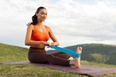 Dağlarda fitness minderinde otururken bacak kaslarını güçlendirmek için elastik bantla egzersiz yapan mutlu zayıf kadın.