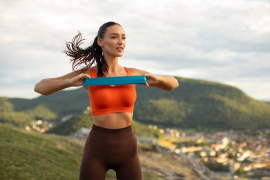 Formda genç bir kadın kol kaslarını spor bandıyla çalıştırıyor. Açık havada, dağın tepesinde duruyor, temiz havanın ve doğanın tadını çıkarıyor.