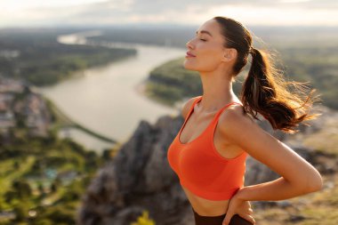 Genç kadın dağlarda antrenman yapıyor, gözleri kapalı nefes alıyor, nehir manzarası ile yoga yapıyor, boş alan arıyor.