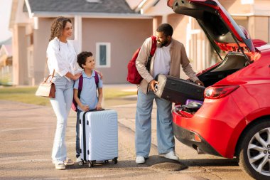 Baba bagajlarını arabaya koyuyor, tatile hazırlanıyor, anne ve oğlu dışarıda otomobilin yanında duruyor.