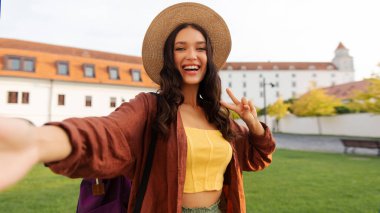 Pozitif genç bayan hasır şapka takıyor ve gülümsüyor V işaretini gösteriyor dışarıda selfie çekiyor, şehirde yürüyor ve geziyor.