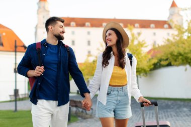 Genç mutlu çift valizleriyle kasabada yürüyor, sırt çantaları takıyor, el ele tutuşuyor ve yolculuğun keyfini çıkarırken birbirlerine gülümsüyorlar.