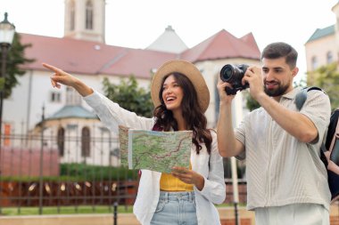 Mutlu genç turistler, harita kullanarak şehri keşfediyor ve kamerayla fotoğraf çekiyor, kadın işaret ediyor ve gülümsüyor.