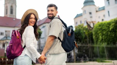 Mutlu Avrupalı iki turist yürüyor, el ele tutuşuyor ve kameraya gülümsüyor, tatillerinde yeni bir şehri keşfediyorlar.