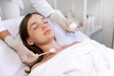 Eldivenli güzellik uzmanı fırça tutuyor ve güzellik salonundaki genç müşterinin yüzüne ve boynuna maske takıyor.