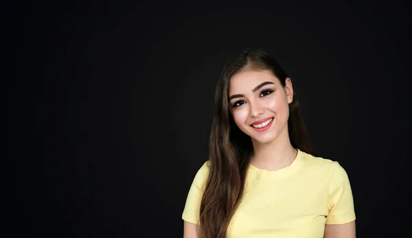 Uzun siyah saçlı genç Asyalı kız, dişleri geniş bir şekilde gülümsüyor, kameraya bakıyor, başı eğik, siyah arka planda siyah tişört giyiyor ve fotokopi çekiyor..