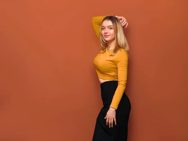 Büyük göğüslü güzel sarışın kızın yan görüntüsü, eli başının arkasında, kameraya bakıyor, sarı bluz ve turuncu arka planda siyah uzun etek giyiyor..