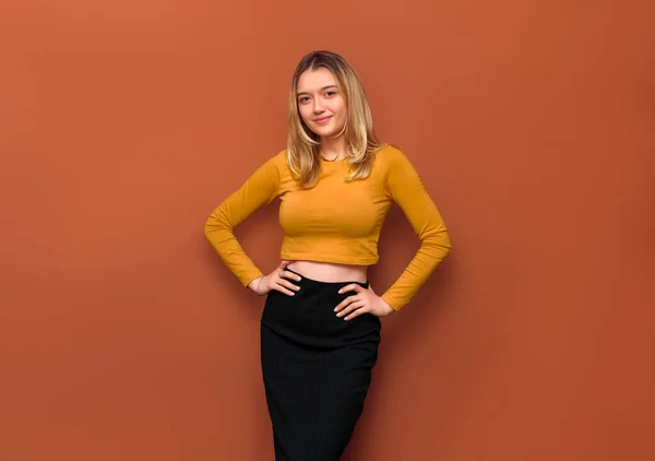 Portrait of beautiful young girl with large breasts, hands on hips, smiling looking at camera, wearing yellow blouse and black long skirt on orange solid background.