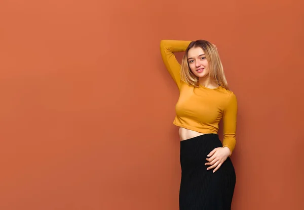 Portrait of beautiful posing girl with big breasts, holding her hand behind head, looking at camera, standing in yellow blouse and black long skirt on orange solid background with copy space.