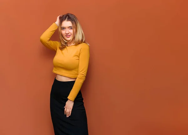 Portrait of beautiful posing blonde girl with big breasts, holding her hand on head, looking at camera, in yellow blouse and black long skirt on orange solid background with copy space.