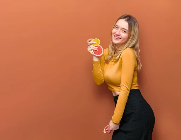 Genç, çekici bir kadın, elinde greyfurt ve portakal dilimleri tutarak poz veriyor, kameraya gülüyor, sarı bluz giyiyor ve vanilya arka planında siyah bir taban ve fotokopi alanı var..