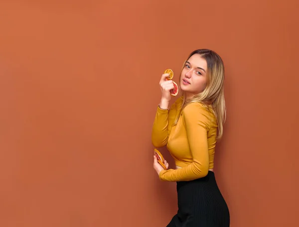Genç ve çekici bir kız ellerinde greyfurt ve portakal dilimleri tutuyor, kameraya bakıyor, yanlamasına duruyor, sarı bluz içinde ve vanilya arka planında siyah tabanlı..