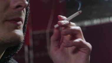 Adam sigara içiyor, duman üflüyor, dudaklarını yalıyor, aşırı yakın plan, yan görüş..