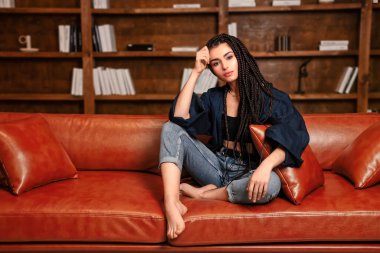 Rastalı kız kitaplığın önündeki koltukta oturuyor. Kafası elinde dinleniyor ve fotokopi makinesiyle kameraya bakıyor..