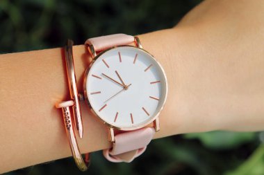 Beyaz yüzlü şık bir kol saati, gül sarısı keçeli kalemler ve gül sarısı bileklik takan yumuşak pembe deri bir kayış. Bulanık yeşillik taze, zarif bir dokunuş ekler..