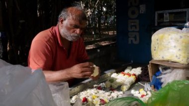 Bangalore, Hindistan-12 Şubat 2023: Yaşlı bir adam güneşli bir günde yol kenarındaki bir çiçekçiye çiçek dizerek çelenk yapıyor