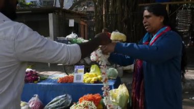 Bangalore,India-February 12,2023:An Indian woman measures and sells a crossandra and jasmine garland at a roadside flower shop during a sunny day