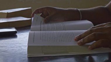 Yakın plan çekimde, masada kitap okurken sayfaları çeviren bir adam görülüyor. Adam kitap okuyor.