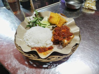 Baharatlı Ayam Penyet (Ezilmiş Tavuk), pilav, kızarmış tempeh, tofu, marul ve salatalık, beyaz bir tabakta servis edilir.