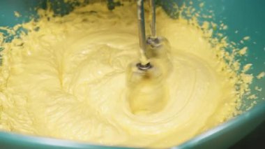 Mangırı bir kasede karıştırıyor, el çırpıcısı kullanıyor, yakınlaştırıyor.